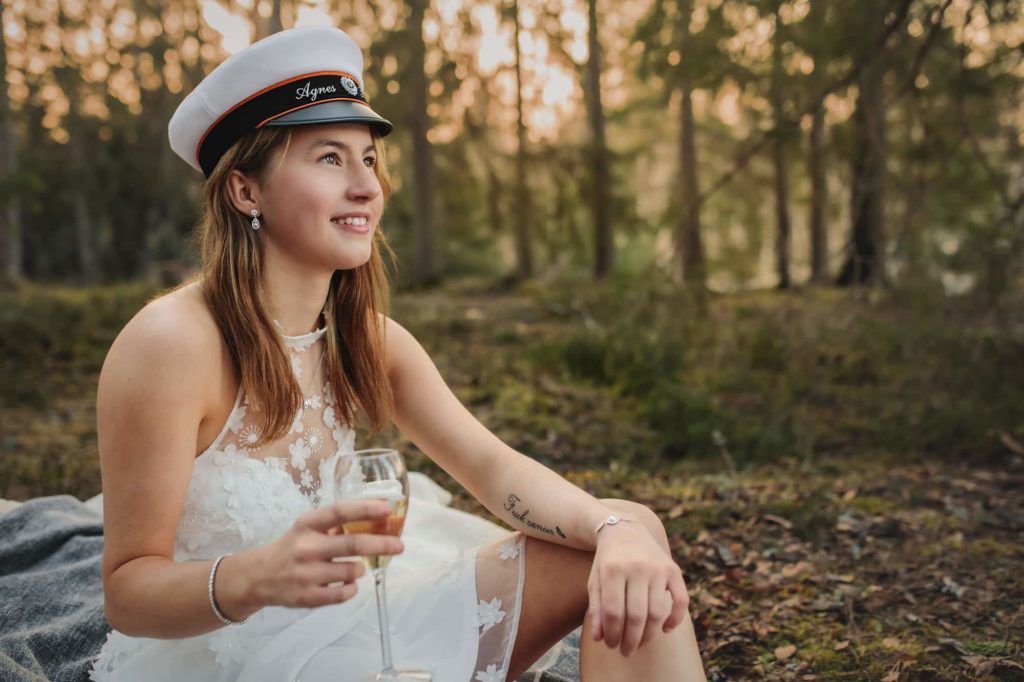 Studentfotografering - Ung kvinna sittandes i skogen med ett glas champagne. Iklädd studenthatt och vit klänning.