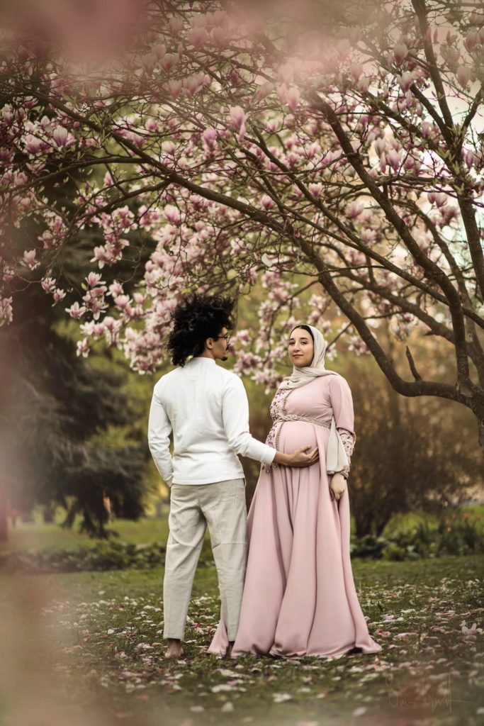 Gravid kvinna i rosa gravidklänning och en man i ljusa kläder, med ryggen mot fotografen, som håller armen under hennes mage.