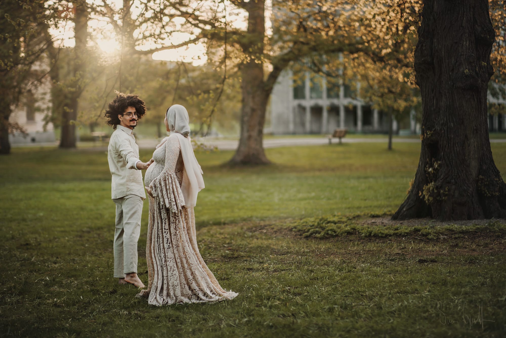 Gravid kvinna i profil med beige gravidklänning. Hennes man, också i beige kläder, håller hennes hand. Solen strålar i bakgrunden i Vasaparken i Västerås .