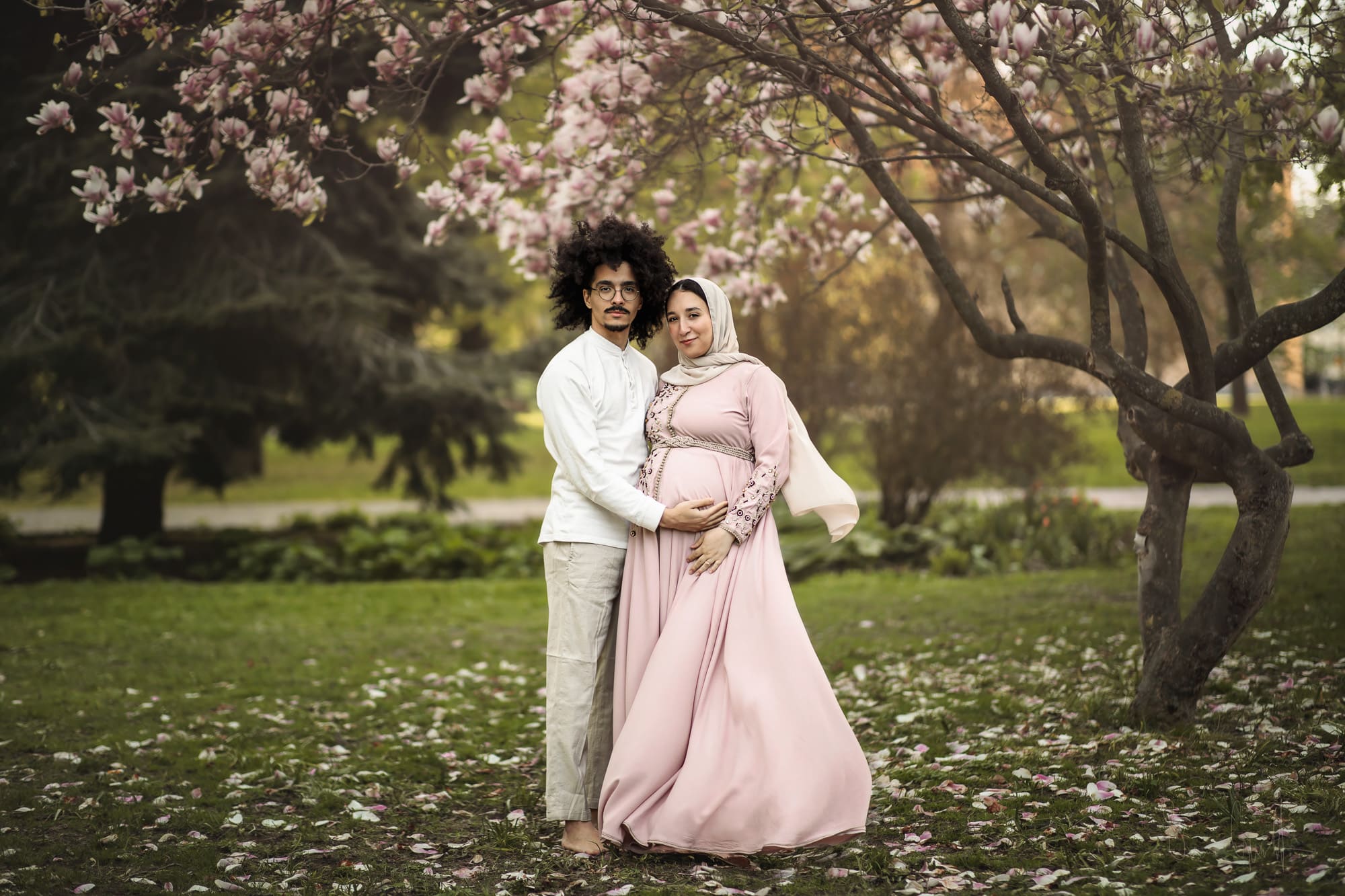Gravid kvinna i rosa gravidklänning och en man i ljusa kläder som håller armen under hennes mage. Vid en gravidfotografering under ett magnoliaträd i Västerås.