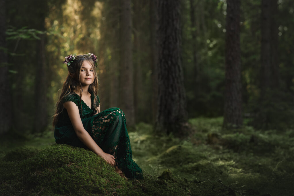 Sagolikt fotografi på en flicka med alvöron och blommor i håret. Sitte spå en mossig sten i en skog med solen glittrandes i bakgrunden