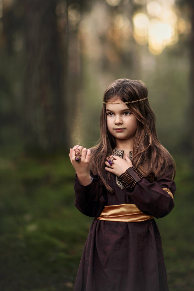 flicka i skogen som har på sig en brun tunika med ett guldfärgat barn i midjan och en guldtråd runt huvudet. ett magiskt glitter sprids från hennes fingrar.