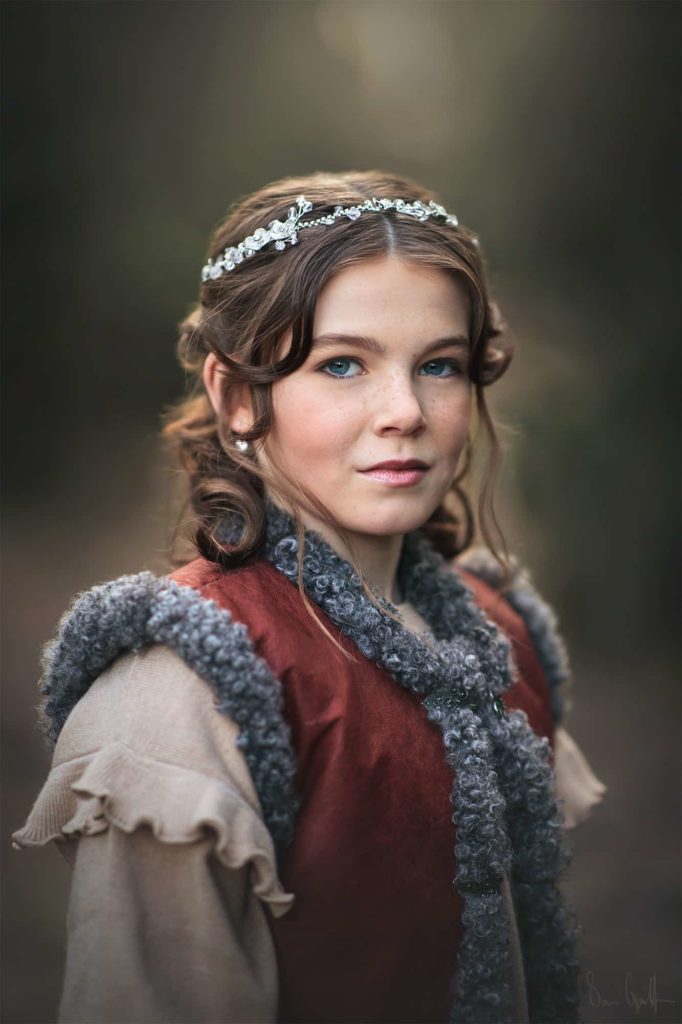 ung tjej i skogen med uppsatt hår och ett glittrande hårsmycke. Hon bär en röd väst med lurvig grå kant.