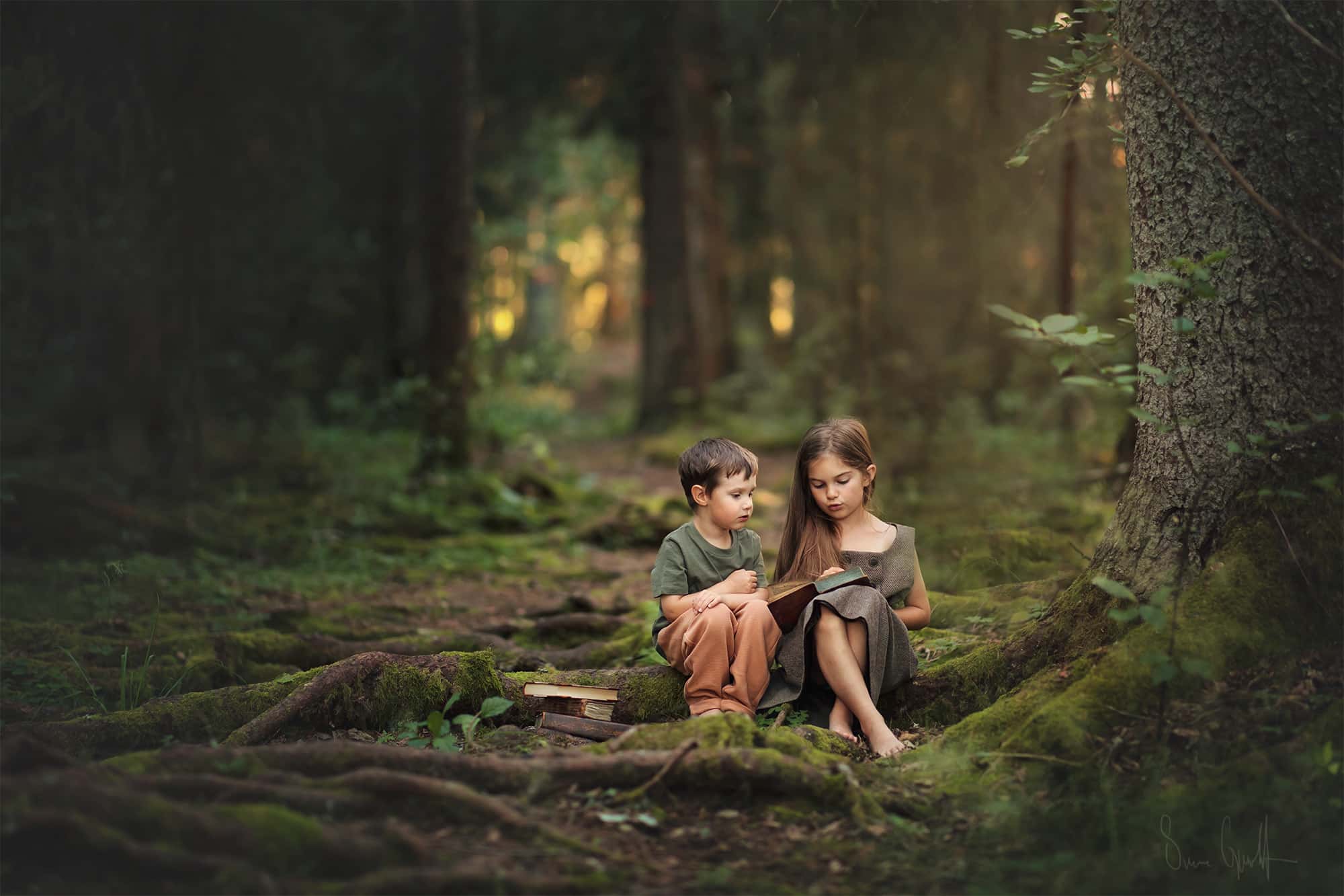 två barn sitter på en stock i skogen. Marken är full av rötter täckta av mossa. Flickan har en beige klänning och pojken en grön tröja och rostfärgade byxor.