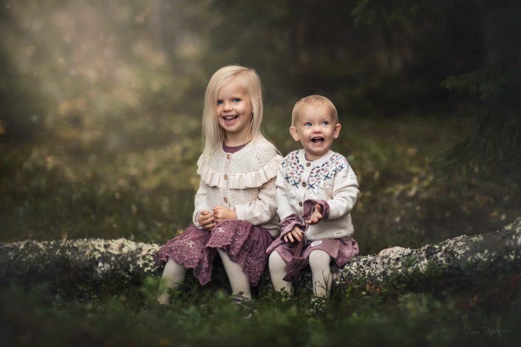 Två små tjejer i lila klänningar och ljusa koftor sitter på en stock i skogen