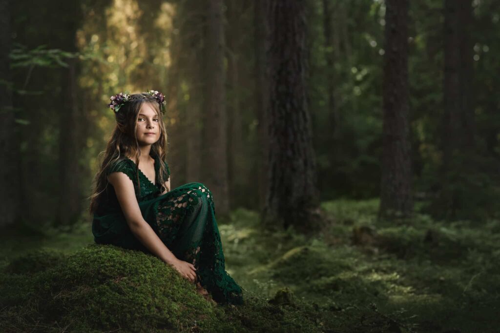 sagolikt foto på flicka med långt brunt hår, grön klänning och blommor i håret, sittandes på en mosstäck sten